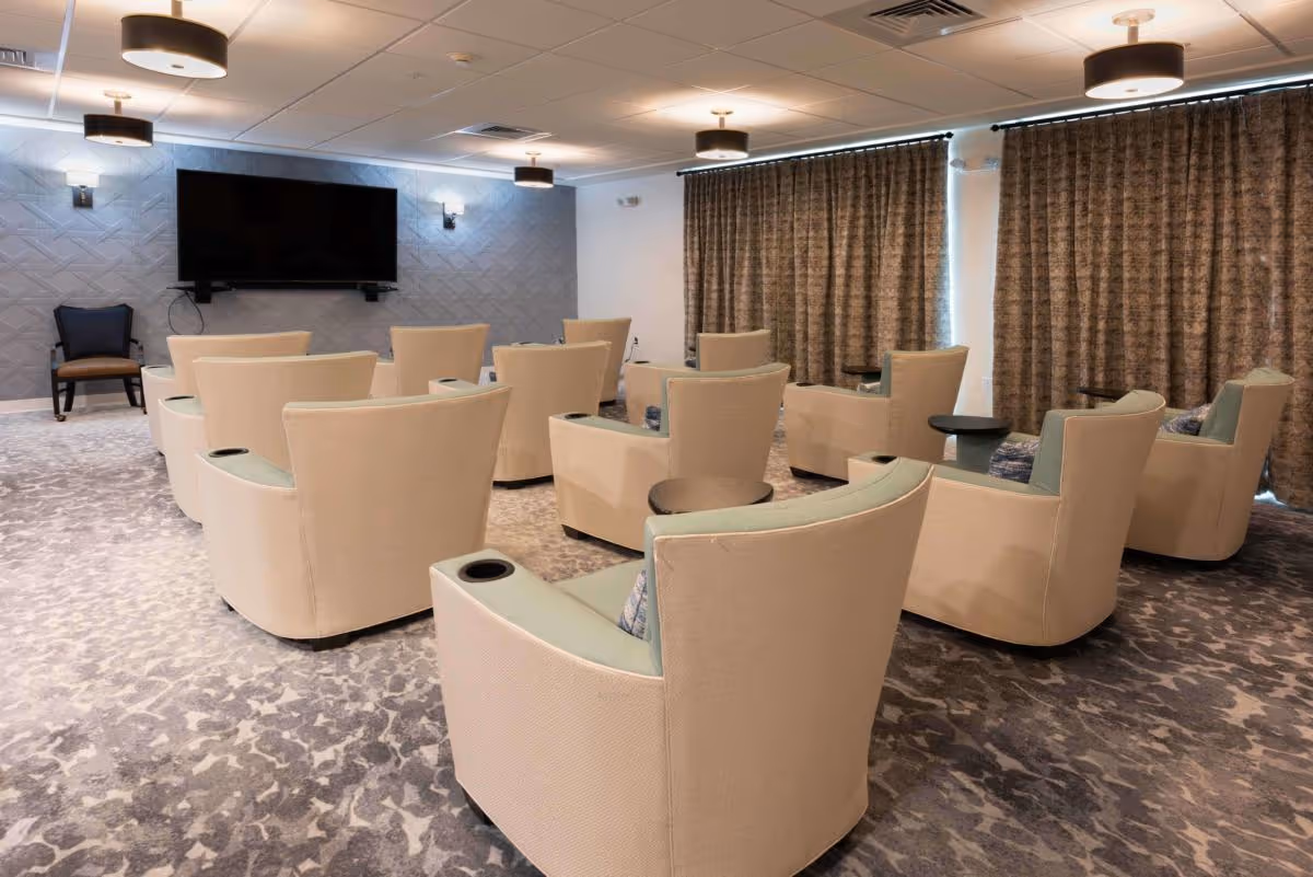 A small theater-style common room with rows of beige armchairs facing a wall-mounted flat-screen TV.