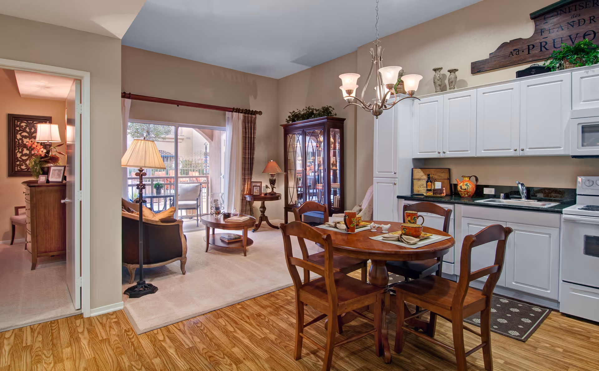 Interior view of a senior living facility showing a combined kitchen and dining area with wooden table and chairs set for two. The kitchen has white cabinets, a sink, and a stove. Adjacent to the kitchen is a living room with a sofa, coffee table, floor lamp, and a glass cabinet. A sliding glass door leads to an outdoor patio with seating. A bedroom is partially visible through an open door on the left.