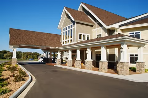 Exterior view of a senior living facility building with a covered entrance supported by stone pillars, a paved driveway, and landscaped areas with shrubs under a clear blue sky.