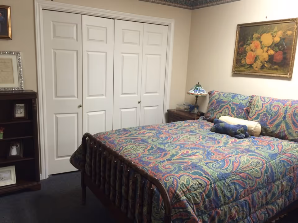 A bedroom with a wooden bed covered in a colorful paisley bedspread, white double closet doors, a nightstand with a lamp, bookshelf, and a floral painting on the wall.