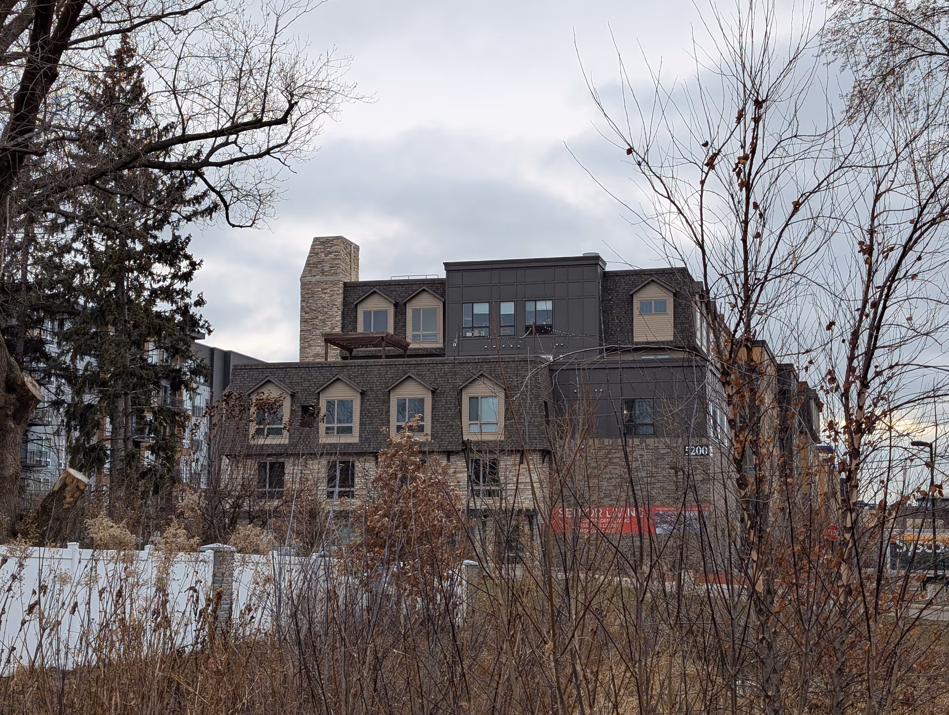 Exterior view of a multi-story senior living facility building with a stone and dark panel facade, surrounded by leafless trees and shrubs under a cloudy sky.