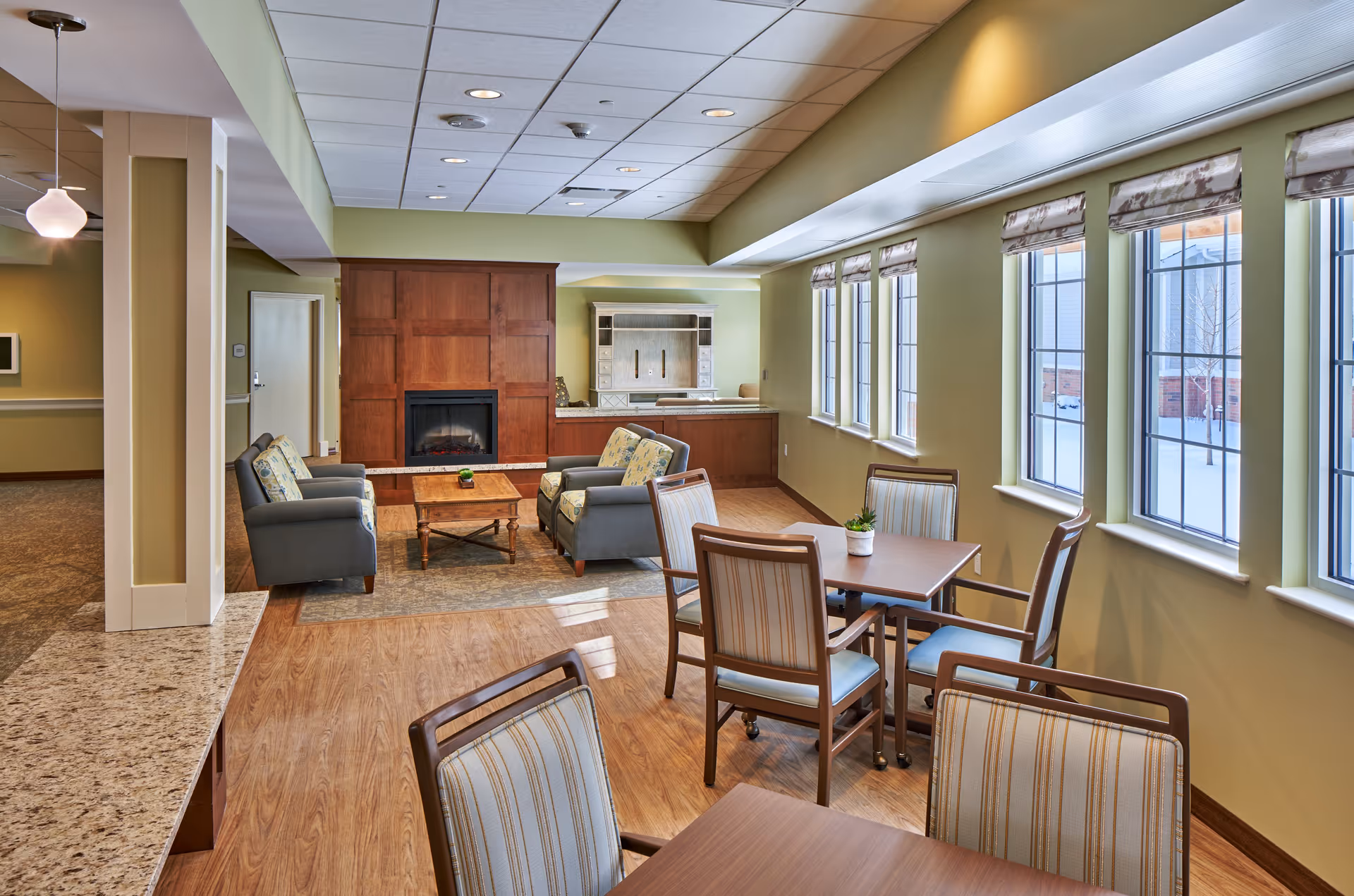 A bright and spacious common area in a senior living facility featuring several tables with chairs near large windows, a seating area with armchairs and a coffee table in front of a wooden fireplace, and a countertop with pendant lighting on the left side.