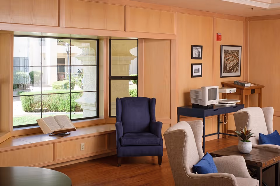A cozy sitting area in a senior living facility with beige armchairs featuring blue cushions, a dark blue wingback chair, a wooden coffee table with a small plant, and a large window with a view of greenery outside. There is a black table against the wall holding an old computer monitor and keyboard, and framed pictures hanging on the wood-paneled wall.
