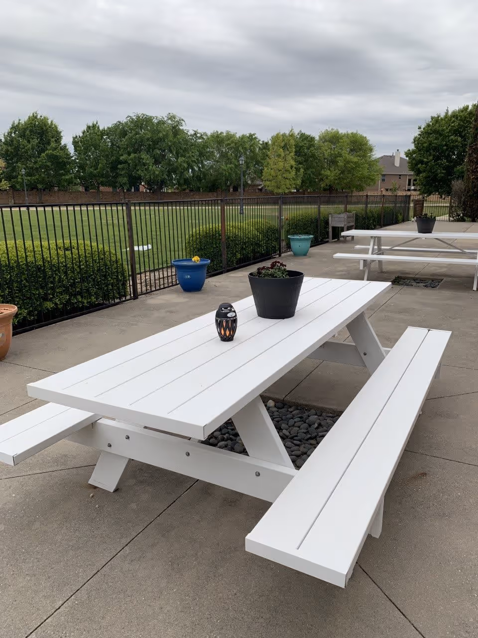 Outdoor patio area with white picnic tables on a concrete surface. Each table has a black flower pot with plants and a small decorative lantern. The area is fenced with black metal fencing, and there are green bushes and trees in the background under a cloudy sky.