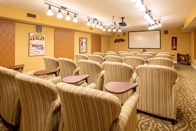 Small screening room with rows of striped upholstered armchairs each with a small attached table facing a projection screen and movie posters on yellow walls.