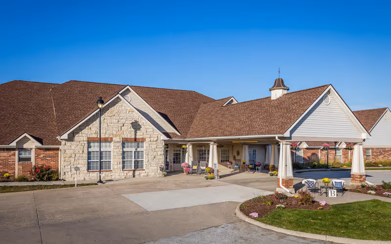 Exterior view of a senior living facility building with a covered entrance, stone and brick facade, and a clear blue sky above. There are some small landscaped areas with flowers and a paved driveway in front.