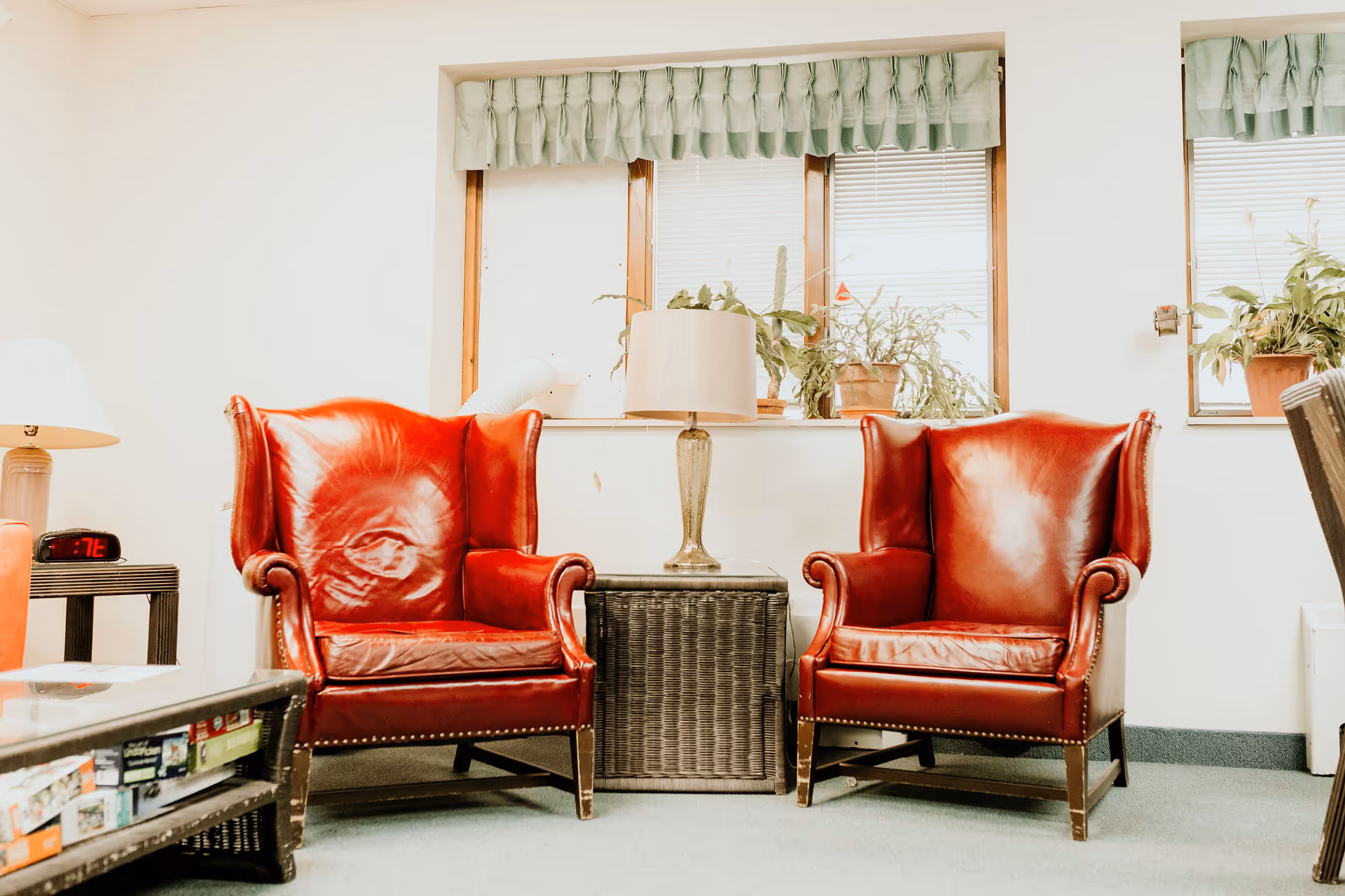 Two red leather wingback chairs with nailhead trim are positioned on either side of a wicker side table with a lamp on it. Behind the chairs is a window with green valances and several potted plants on the windowsill. A coffee table with board games underneath is partially visible in the foreground.