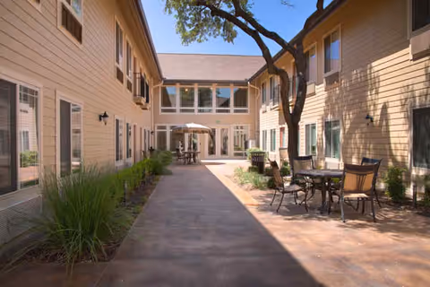 Outdoor courtyard area between two beige buildings with multiple windows. There are several tables and chairs set up along the right side under a tree, and an umbrella-covered table is visible in the distance. The sky is clear and blue.