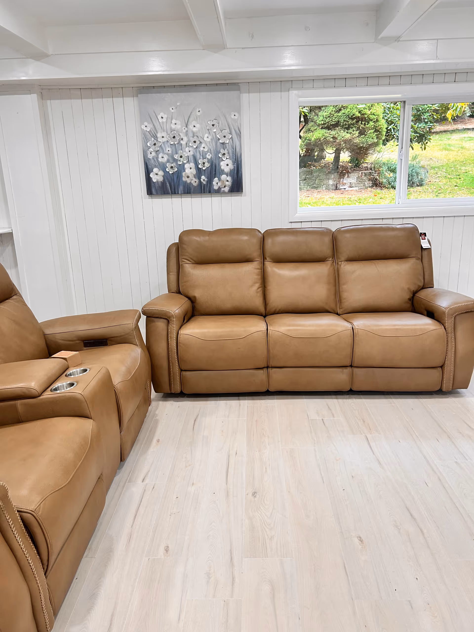 A cozy living room area with a tan leather three-seater sofa and a matching recliner chair with cup holders. The walls are white with vertical paneling, and there is a window showing greenery outside. A painting of white flowers on a gray background hangs on the wall above the sofa.