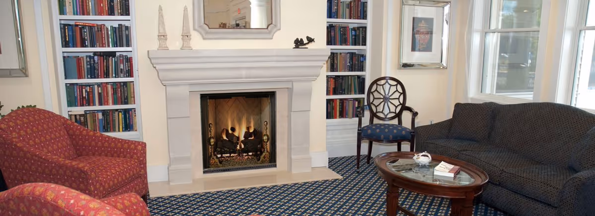 Cozy living room with a lit fireplace, two red patterned armchairs, a dark blue sofa, a wooden coffee table with books and a decorative item, a wooden chair with a blue cushion, built-in bookshelves filled with books, and large windows letting in natural light.