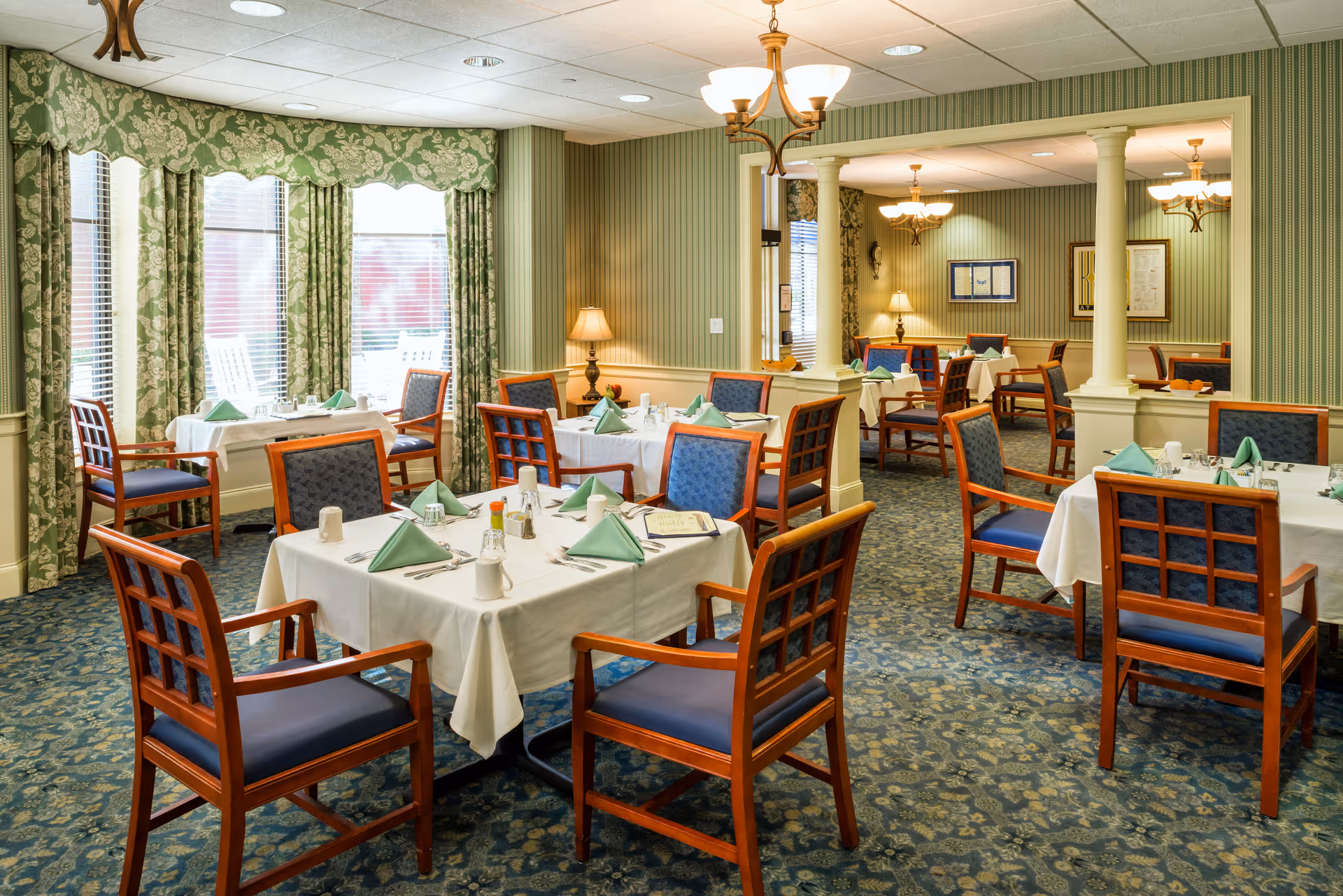 A well-lit dining room with several square tables covered with white tablecloths, each set with green folded napkins, glasses, and condiments. The room features wooden chairs with blue cushioned seats and backs, green patterned curtains on the windows, and green striped wallpaper. There are chandeliers hanging from the ceiling and a carpeted floor with a floral pattern.