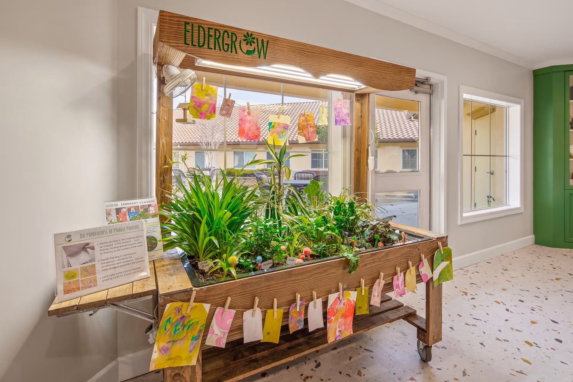 Indoor gardening station labeled 'Eldergrow' with various green plants growing in a wooden planter. Colorful artwork is clipped and hanging around the planter. The station is set against a window with a view of a building with a tiled roof outside. The room has light-colored walls and a terrazzo floor with speckled patterns.