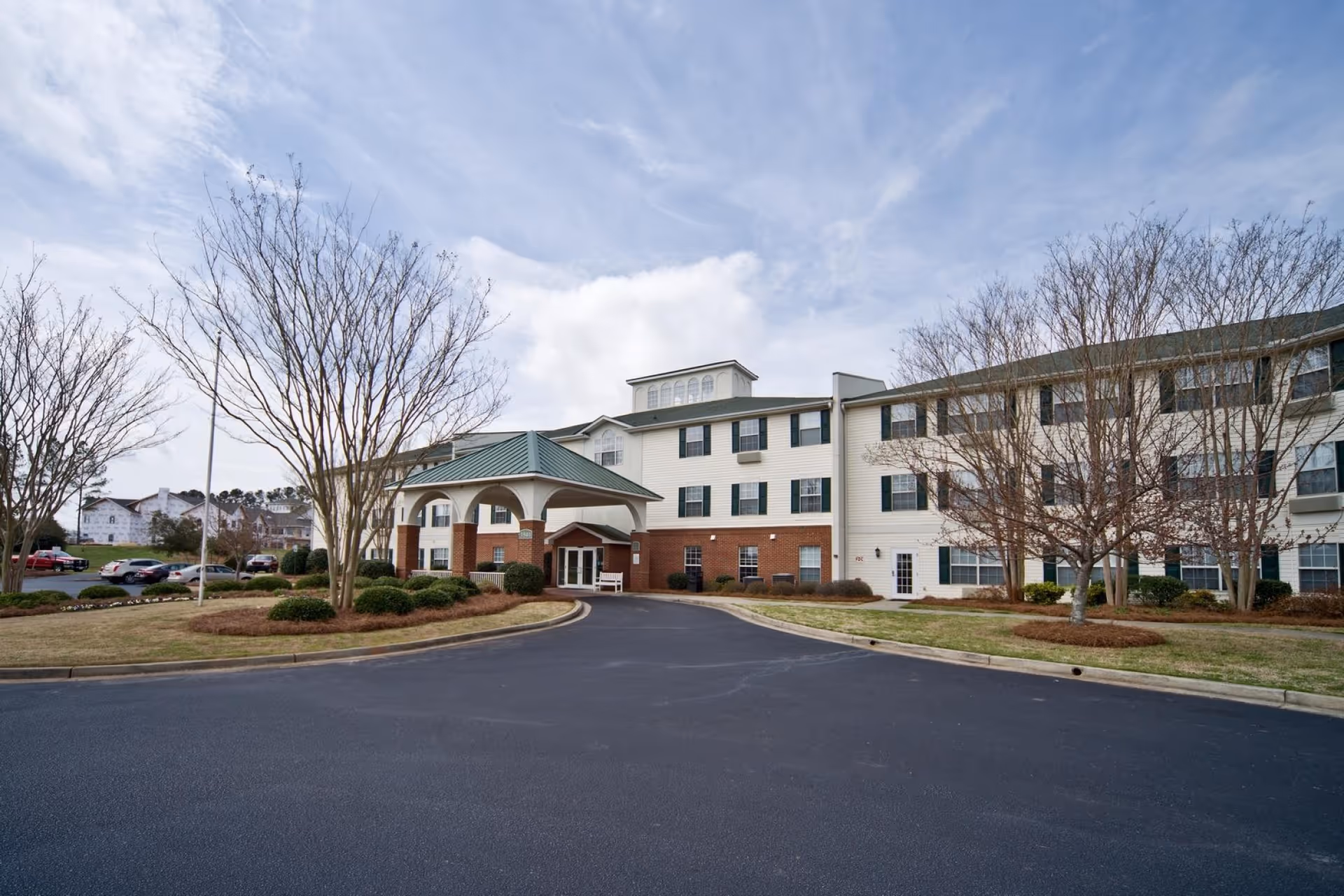 Exterior view of The Legacy of Anderson senior living facility showing a three-story building with a covered entrance, surrounded by leafless trees and a parking area under a partly cloudy sky.