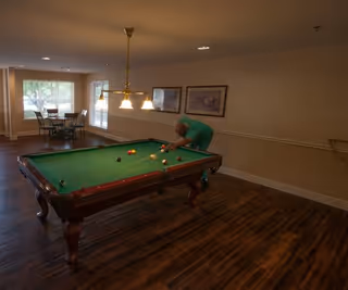 A person playing pool on a billiards table in a room with wooden flooring, beige walls, and framed pictures. There is a hanging light fixture above the pool table and a small dining table with chairs near large windows in the background.