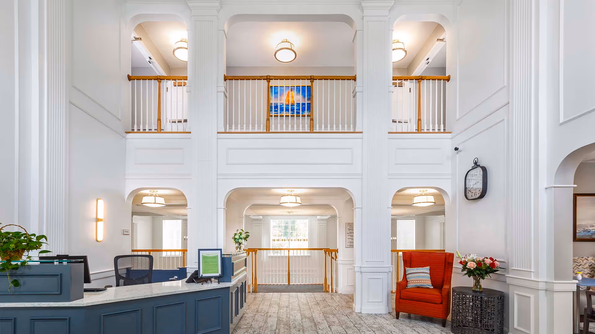 Bright and spacious reception area with white walls and high ceilings featuring large columns. A blue reception desk with office chairs is on the left, and a red armchair with a small table holding a flower vase is on the right. The upper level has a wooden railing and a painting of a sailboat on the wall. A large clock is mounted on the right wall.