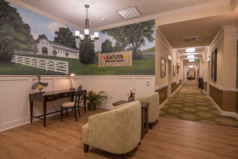Well-lit senior living facility interior with seating and a desk in front of a large mural reading 'SATURN Welcome Center' and a long carpeted corridor.