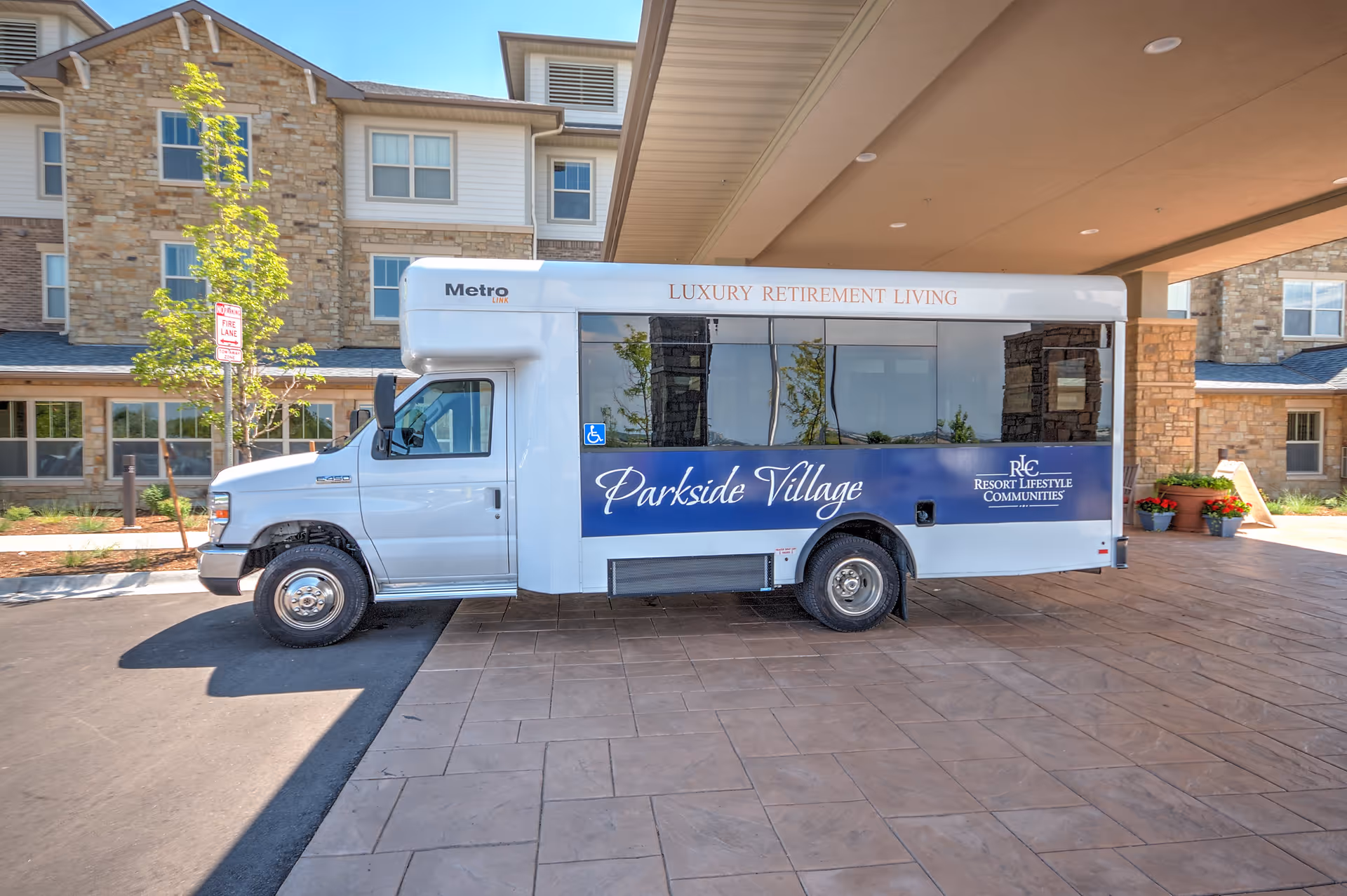 A white shuttle bus parked under a covered driveway in front of a stone and siding building. The bus has signage that reads 'Parkside Village' and 'Luxury Retirement Living' along with the Resort Lifestyle Communities logo. There are small trees and plants around the building.