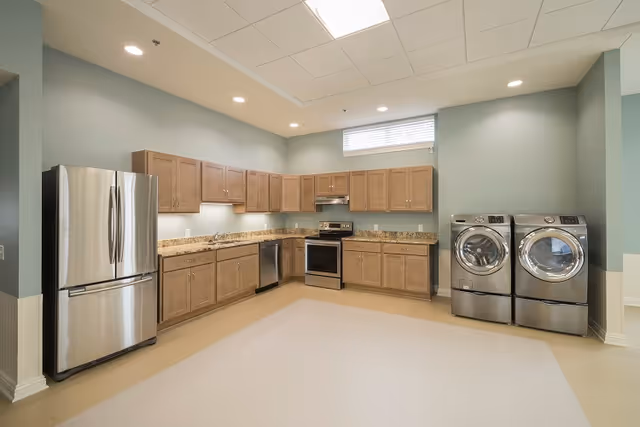 Spacious communal kitchen with stainless steel refrigerator, range and dishwasher, wooden cabinets, granite countertops, and a stacked washer and dryer.