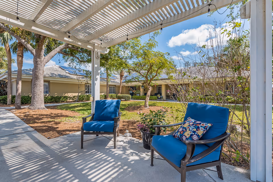 Outdoor seating area at Healthpark Senior Living featuring two blue cushioned chairs under a white pergola with a patterned pillow on one chair. The area is surrounded by trees, plants, and a well-maintained garden with a building in the background under a blue sky with some clouds.