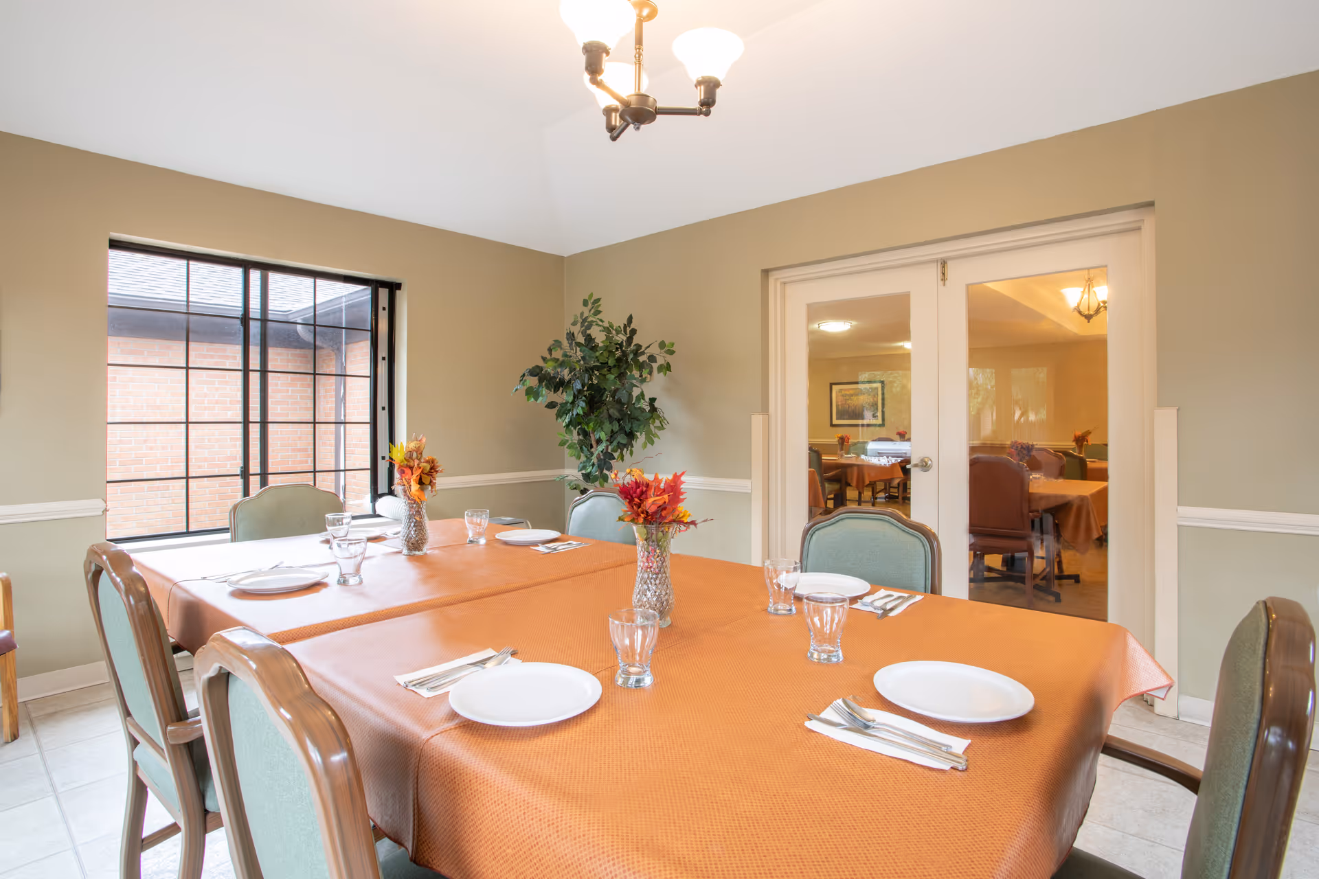 A dining room with a rectangular table covered with an orange tablecloth, set with white plates, glasses, and silverware. There are six chairs around the table, a large window showing a brick exterior wall, a potted plant in the corner, and double glass doors leading to another room with more tables and chairs.