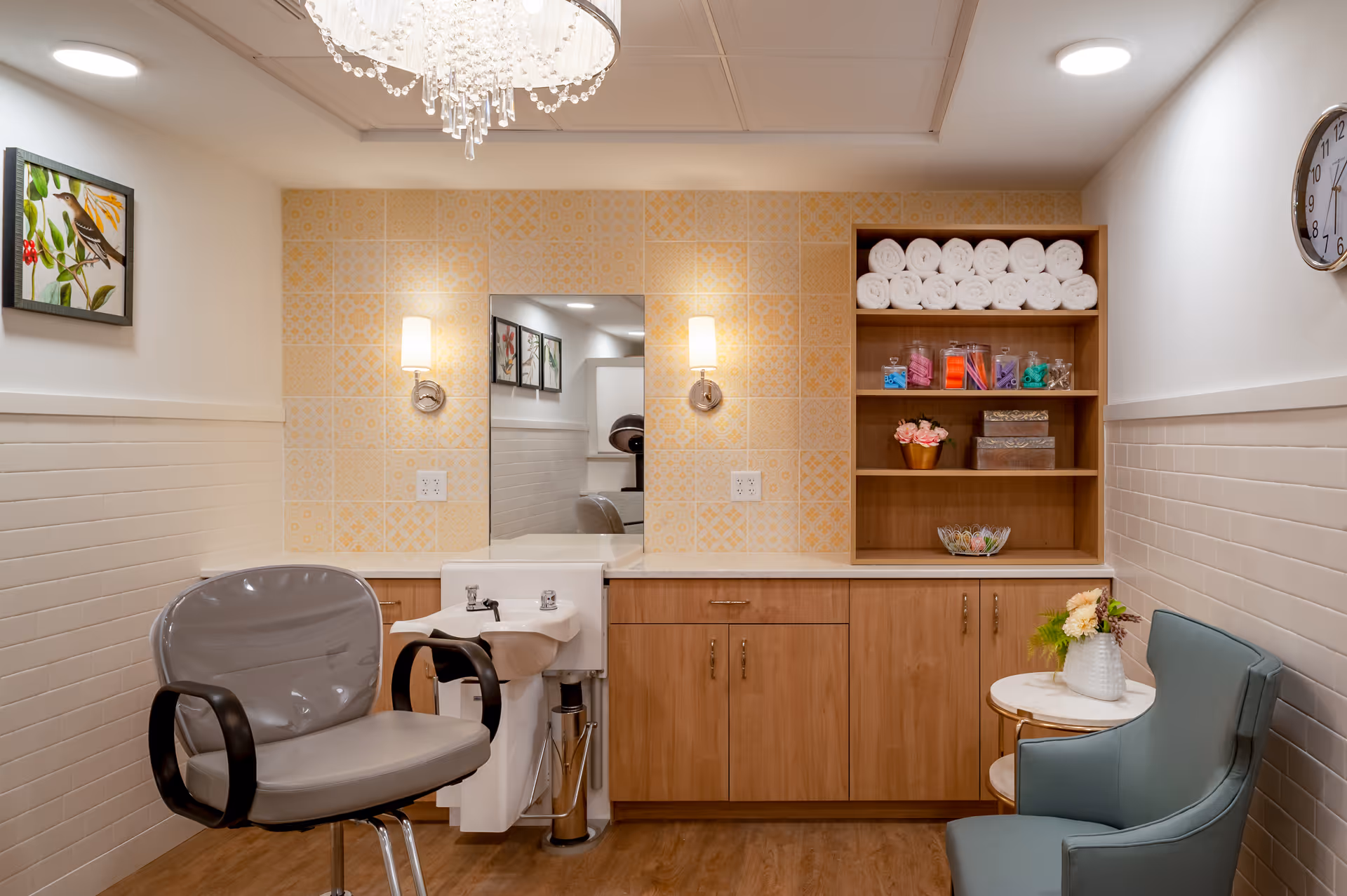 A well-lit salon or grooming area with a gray salon chair in front of a small white sink. The wall behind features yellow patterned tiles, a mirror, and two wall sconces. To the right, there is a wooden cabinet with shelves holding rolled white towels, colorful jars, and decorative items. A small round table with a flower vase and a teal upholstered chair are also visible. A clock hangs on the right wall.