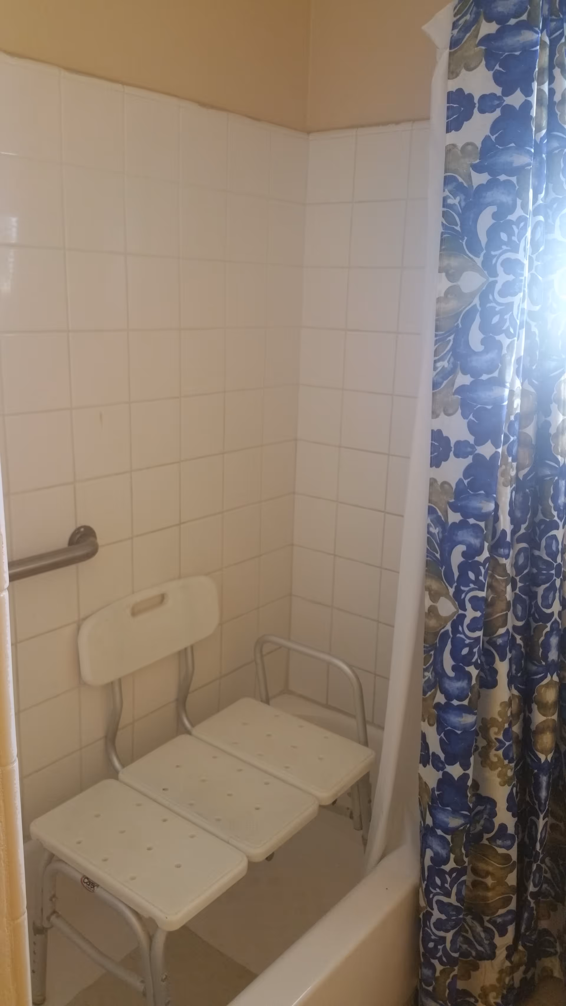 Bathtub/shower with tiled walls, a white shower transfer bench and grab bar, and a blue floral shower curtain.