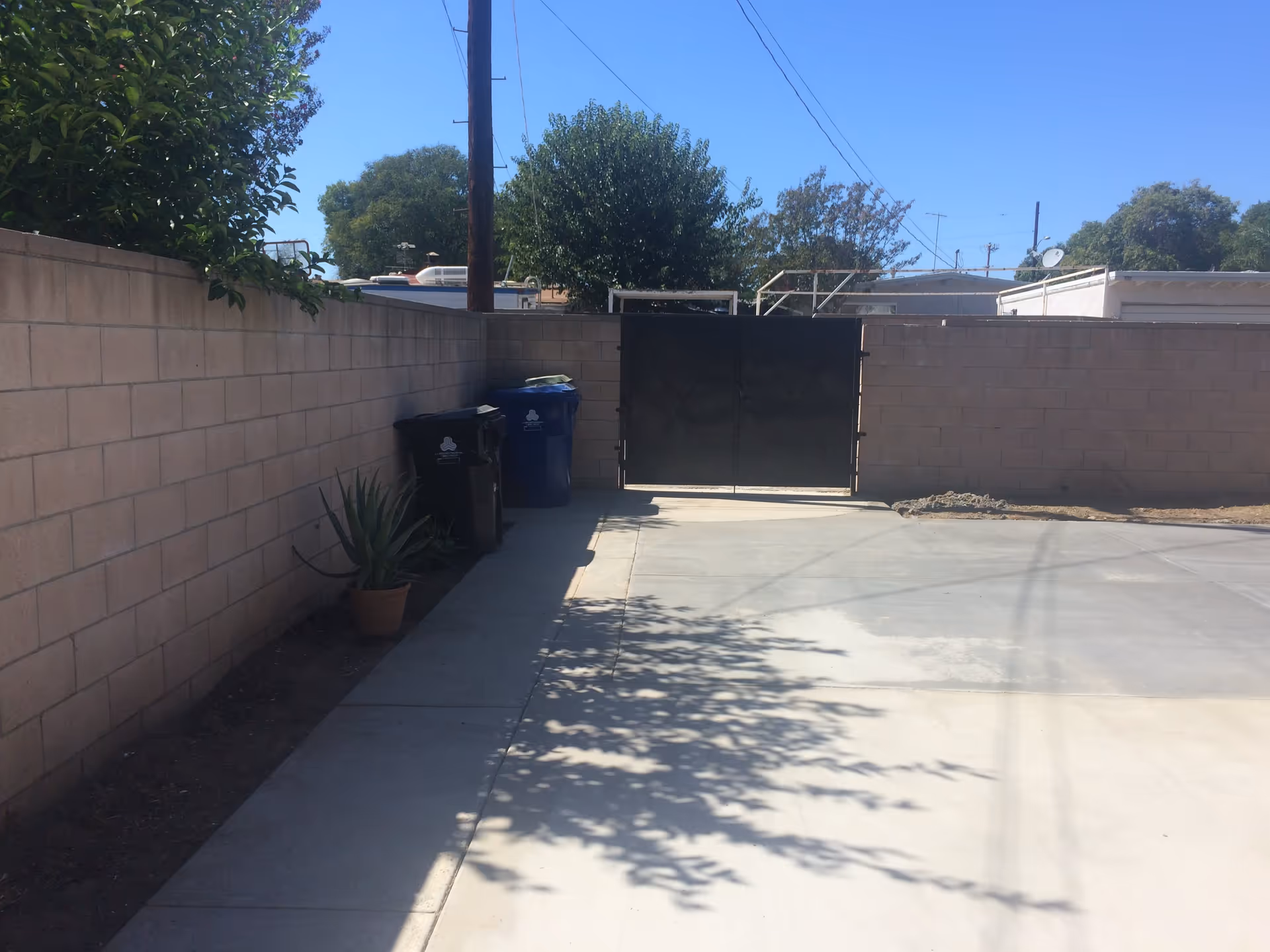 Outdoor area with a concrete pathway and driveway next to a brick wall. There are two trash bins and a potted plant along the wall. A black metal gate is visible at the end of the pathway, with trees and a clear blue sky in the background.