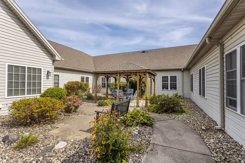 Outdoor courtyard area at Brookdale Grayson View featuring a wooden gazebo with seating underneath, surrounded by bushes, flowers, and a paved walkway. The courtyard is enclosed by single-story buildings with white siding and multiple windows under a partly cloudy sky.