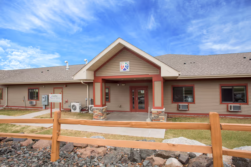 Exterior view of Carefree Living Silver Bay facility showing a single-story building with beige siding and red trim. The entrance has a covered porch with stone pillars and double glass doors. A wooden fence and rocks are in the foreground, with a bright blue sky and scattered clouds above.