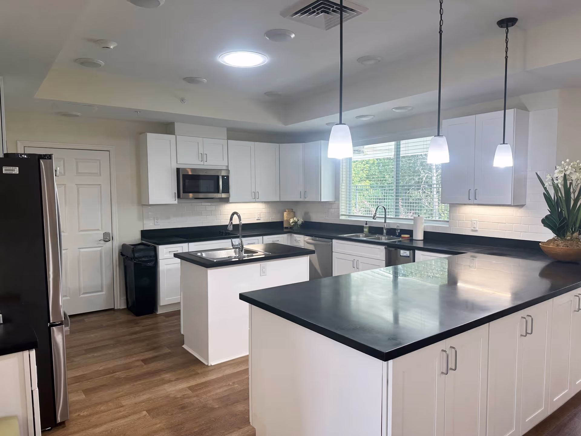 A modern kitchen with white cabinets, black countertops, and wood flooring. The kitchen features a central island with a sink, a stainless steel refrigerator, a built-in microwave above the stove area, and a large window letting in natural light. Three pendant lights hang from the ceiling above the counters.