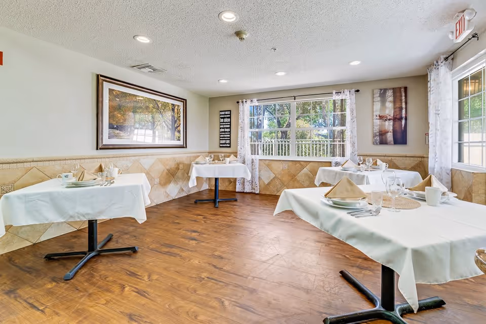 A bright dining room in a senior living facility with four tables covered in white tablecloths, each set with plates, napkins, glasses, and silverware. The room has large windows with sheer curtains allowing natural light to fill the space. The walls are decorated with framed pictures and the floor is wooden.