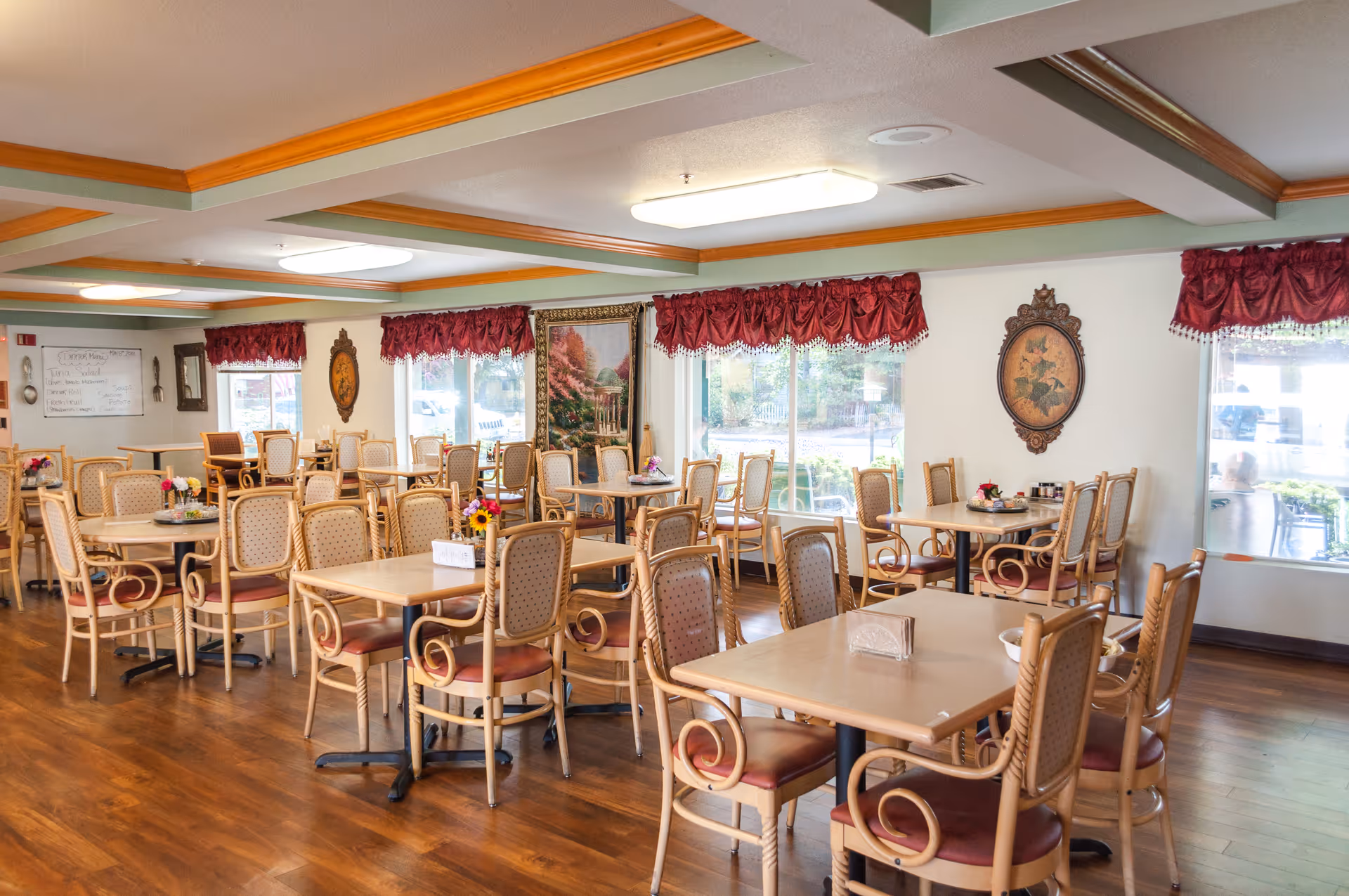 A bright dining room with multiple wooden tables and chairs arranged neatly. The room has large windows with red valance curtains, wooden flooring, and decorative wall art. There is a whiteboard with a menu written on it in the background.