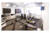A fitness room with exercise equipment including a treadmill, stationary bike, weight bench, and dumbbells. The room has large windows letting in natural light and two large flat-screen TVs mounted on the walls.