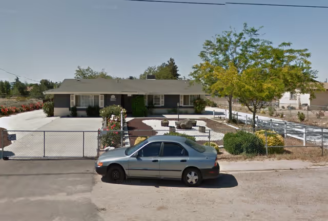 Single-story residential building with a fenced front yard featuring a landscaped garden with rocks and bushes. A silver sedan is parked on the street in front of the property. The sky is clear and blue.