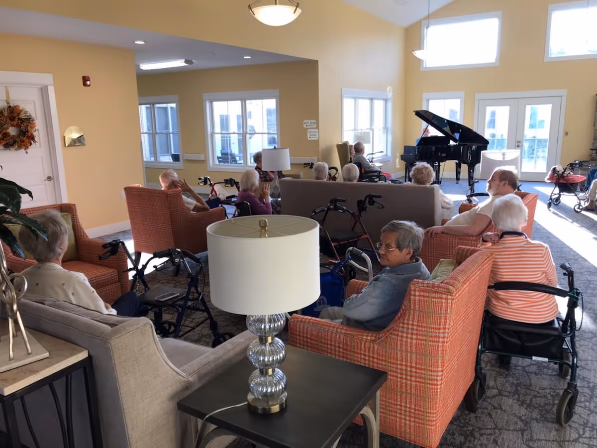 A group of elderly people seated in a living room area with armchairs and sofas, some using walkers, facing a grand piano near large windows and glass doors letting in natural light.