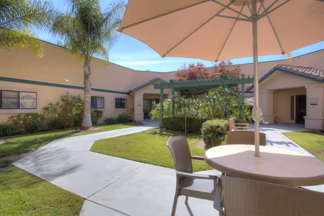 Outdoor courtyard area at Summerfield of Encinitas Memory Care featuring a round table with chairs and a large umbrella, surrounded by a concrete walkway, green grass, palm trees, and a building with beige walls and green trim under a clear blue sky.