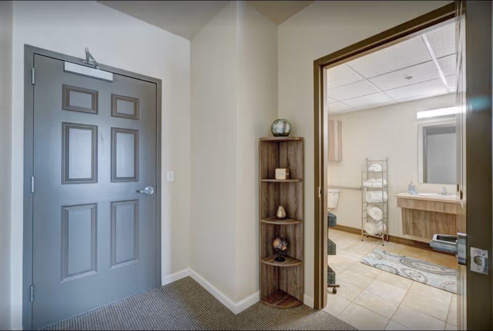 View of an entryway with a closed gray door on the left and an open door on the right leading to a bathroom. The bathroom has a sink with a mirror, a toilet, a towel rack with rolled white towels, and a patterned rug on the tiled floor. A small wooden corner shelf with decorative items is positioned between the two doors.