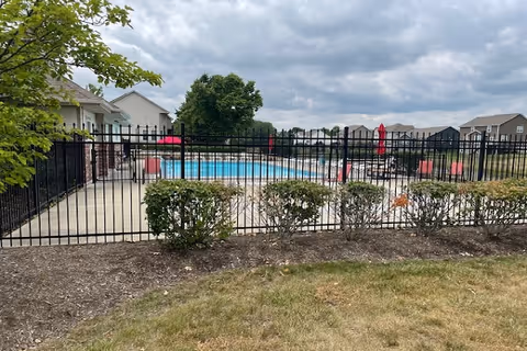 Outdoor swimming pool area enclosed by a black metal fence with bushes in front. There are several red umbrellas and lounge chairs around the pool. Residential buildings and trees are visible in the background under a cloudy sky.