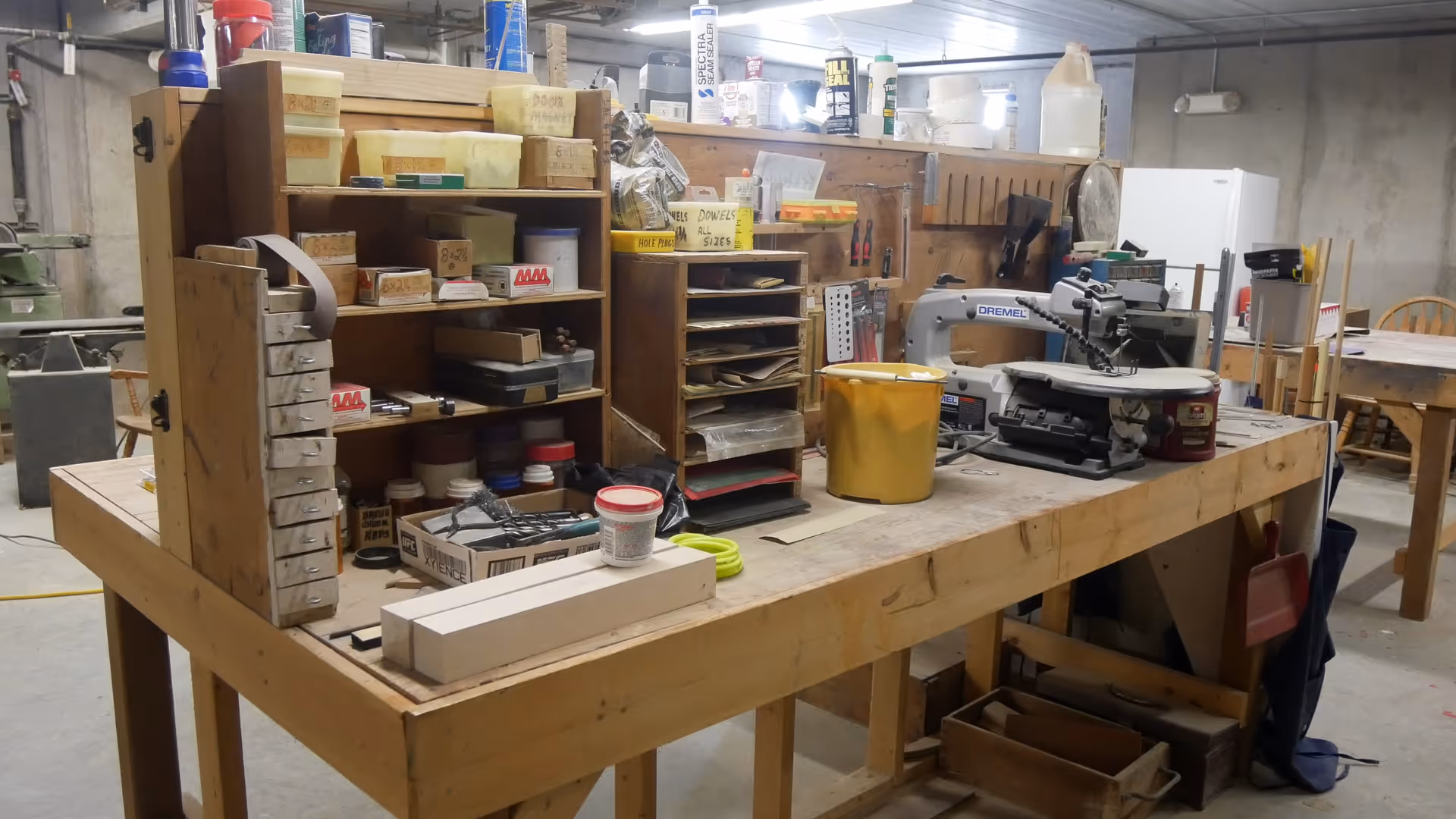 Woodworking workshop interior with a large wooden workbench covered in tools, organizers, and a scroll saw.