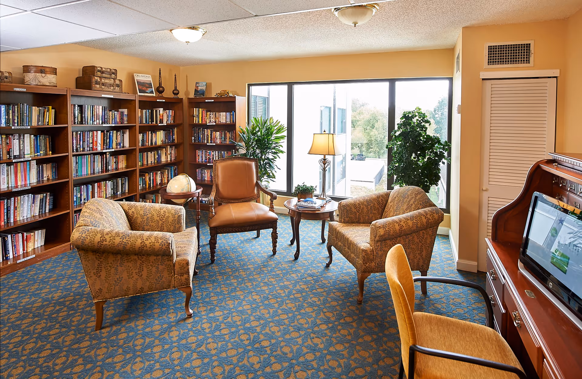 A cozy reading room with three upholstered armchairs and a leather chair arranged around a small wooden table with a lamp. There are bookshelves filled with books along the left wall, a large window letting in natural light, and a wooden desk with a computer on the right side. The room has a patterned blue carpet and beige walls with some plants near the window.