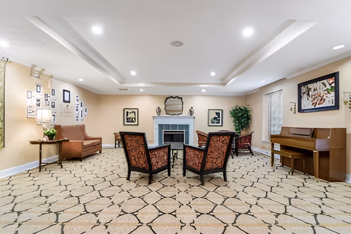 Spacious common living room with chairs arranged around a central fireplace, a piano, and seating along the walls.