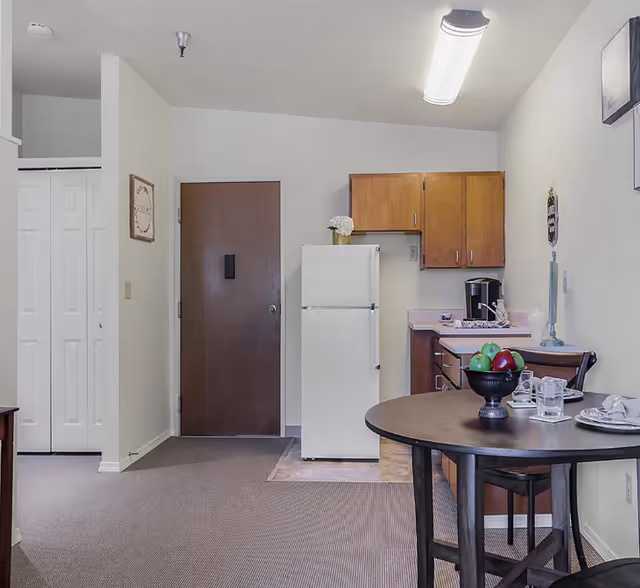 Studio kitchenette and dining area with a white refrigerator, wood cabinets, and a round table set with dishes and a bowl of fruit.