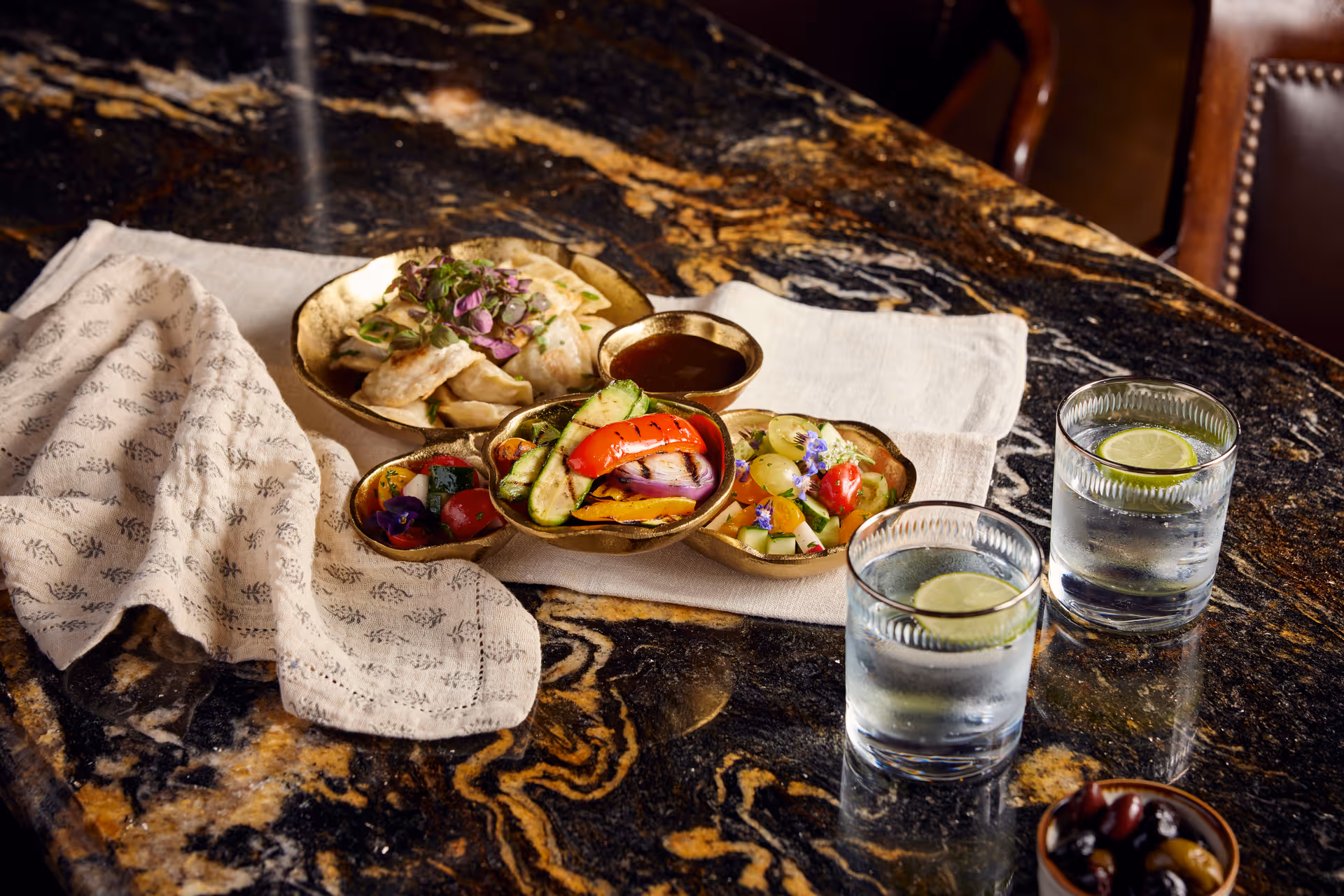A close-up view of a dark marble table set with a beige cloth napkin, a plate of dumplings garnished with herbs, three small bowls containing grilled vegetables, a fresh fruit salad, and a dipping sauce. Two glasses of water with lime slices are also on the table, along with a small bowl of mixed olives.