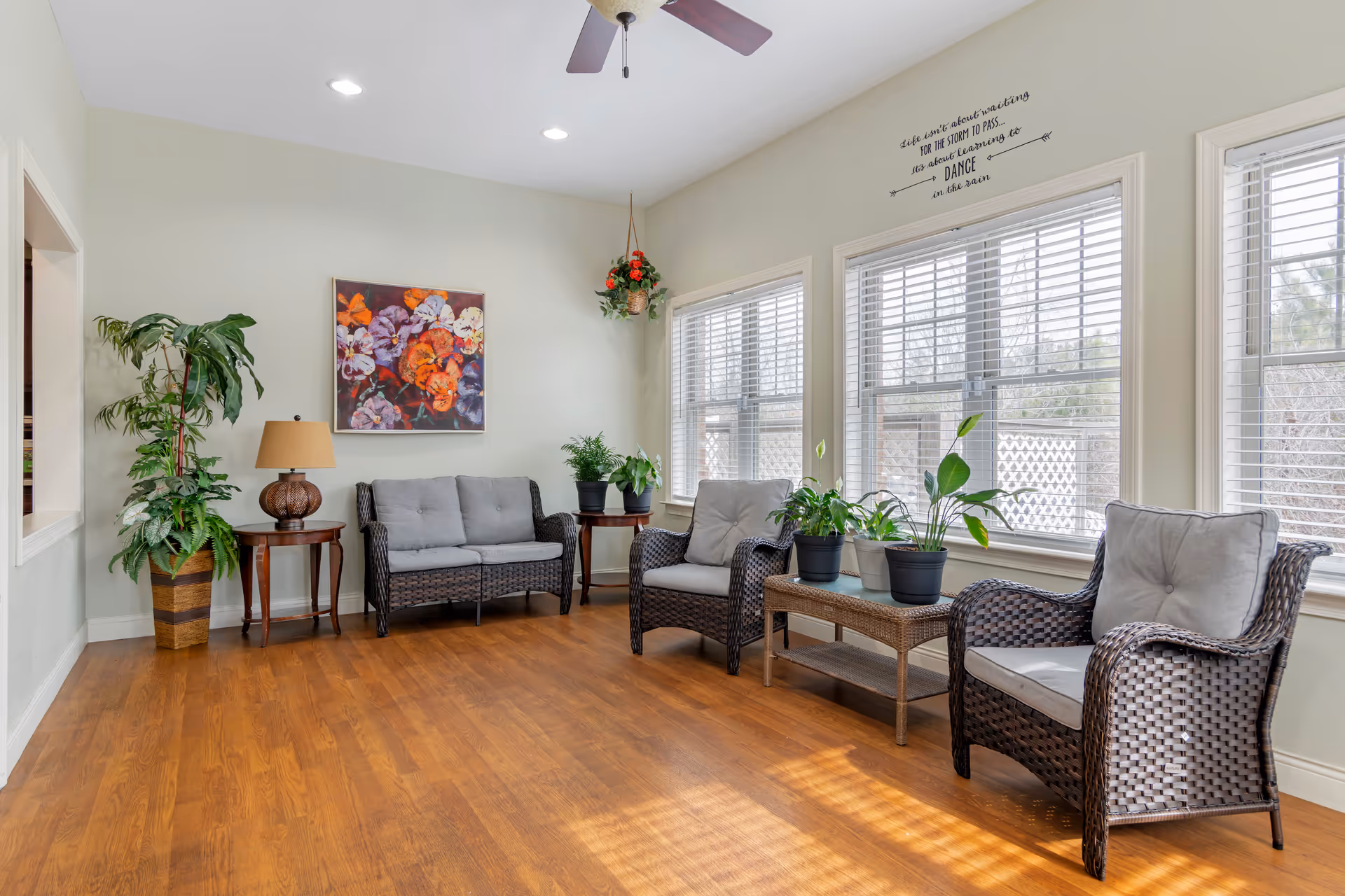 A bright and cozy sitting area with wooden flooring, featuring two cushioned wicker armchairs, a cushioned wicker loveseat, a wicker coffee table with potted plants, and two side tables with a lamp and plants. Large windows with white blinds allow natural light to fill the room. A colorful floral painting hangs on the wall, and a hanging plant is suspended near the windows. Above the windows, a wall decal reads, 'Life isn't about waiting for the storm to pass... it's about learning to dance in the rain.'