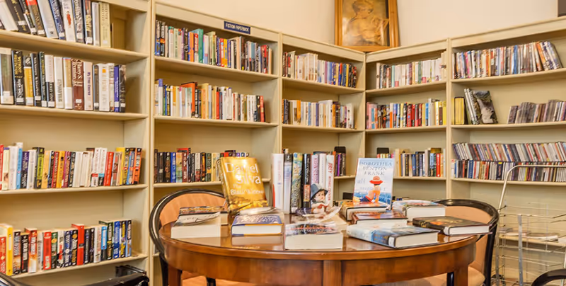 A cozy library room with shelves filled with books and CDs. A round wooden table in the center holds several books and a small figurine. There are chairs around the table and a framed picture on the wall above the shelves.