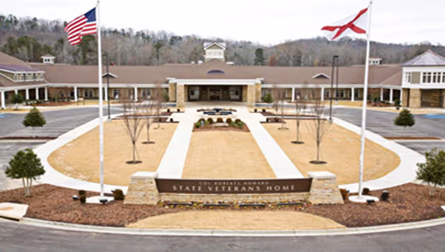 Front exterior view of Colonel Robert L. Howard State Veterans Home with two flagpoles displaying the American flag and the Alabama state flag, a circular driveway, landscaped grounds, and a large building in the background.