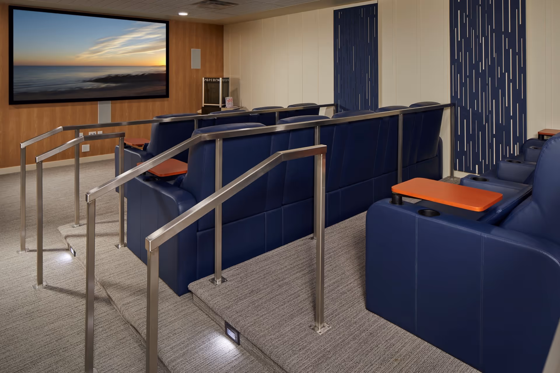 A small private theater room with two rows of blue leather reclining seats, each equipped with orange tray tables and cup holders. The room has carpeted flooring, metal handrails along the steps, and a large screen on the front wall displaying a sunset over the ocean. There is a popcorn machine in the corner.