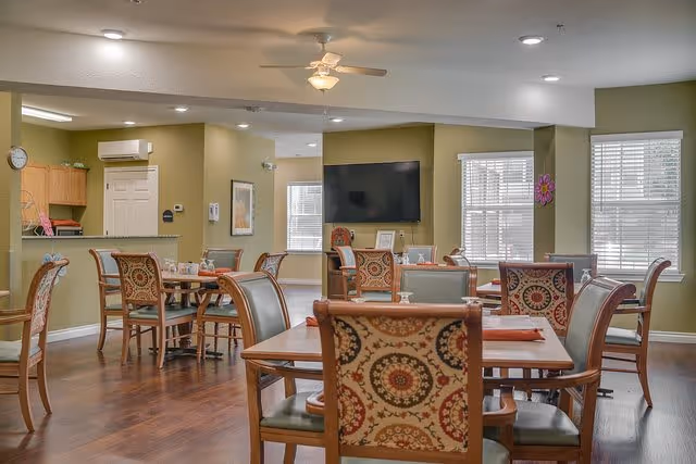 A dining area in a senior living facility with multiple wooden tables and chairs featuring patterned upholstery. The room has green walls, large windows with blinds, a ceiling fan with a light, and a wall-mounted flat screen TV. The floor is wooden, and there is a small kitchen area visible in the background.