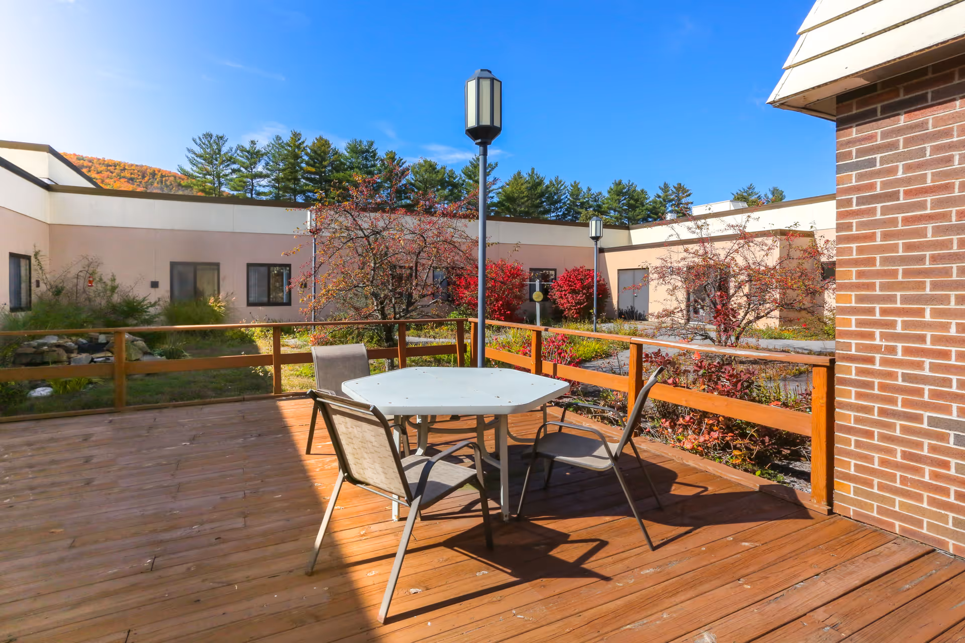 Outdoor wooden deck area with a white hexagonal table and four chairs. The deck is surrounded by a wooden railing and has two tall lamp posts. In the background, there are trees with autumn foliage and a beige building with several windows under a clear blue sky.