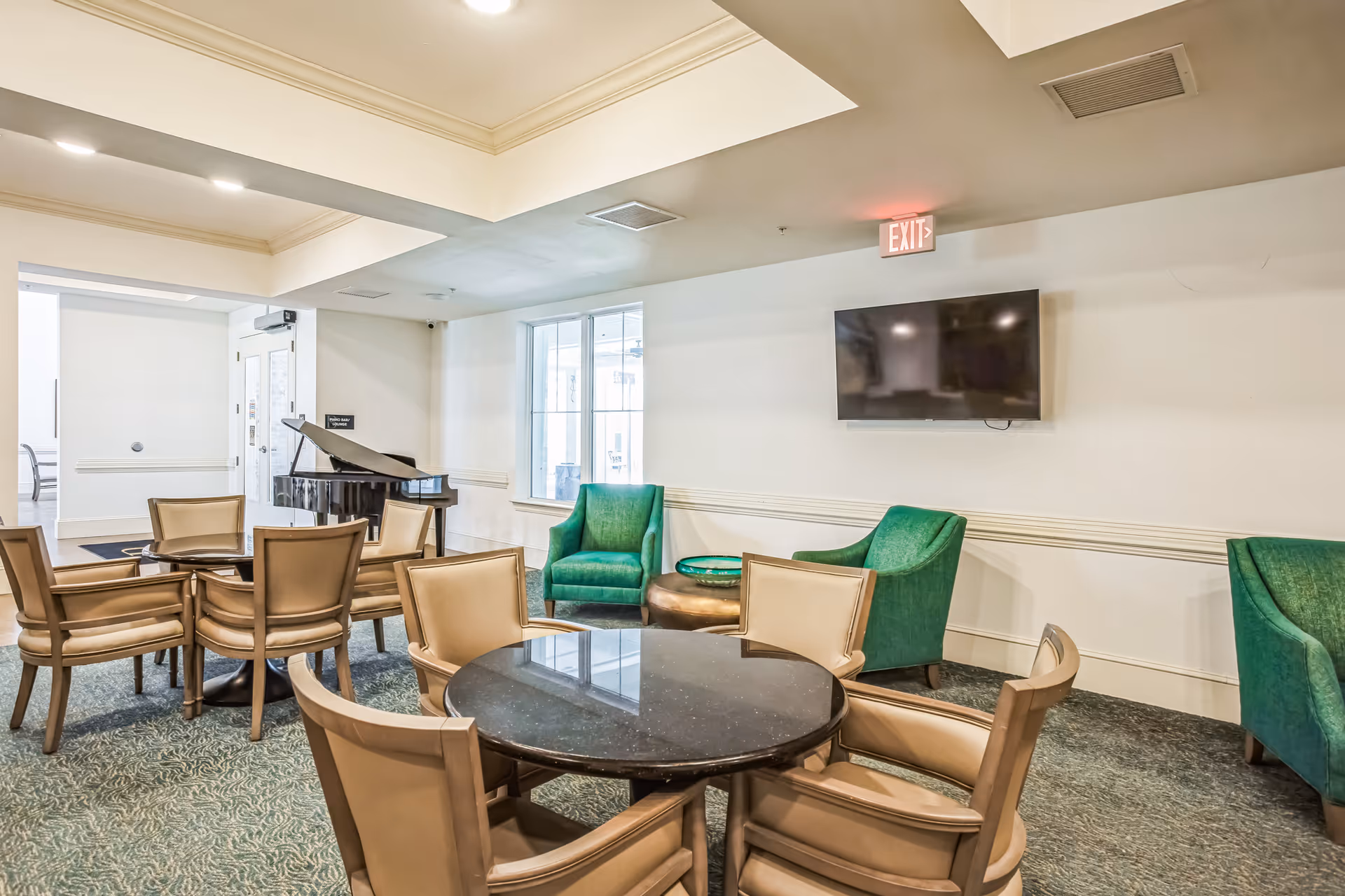 A well-lit common area in The Blake at Colonial Club featuring round tables with beige chairs, green armchairs, a wall-mounted flat screen TV, and a black grand piano near a window and entrance door.